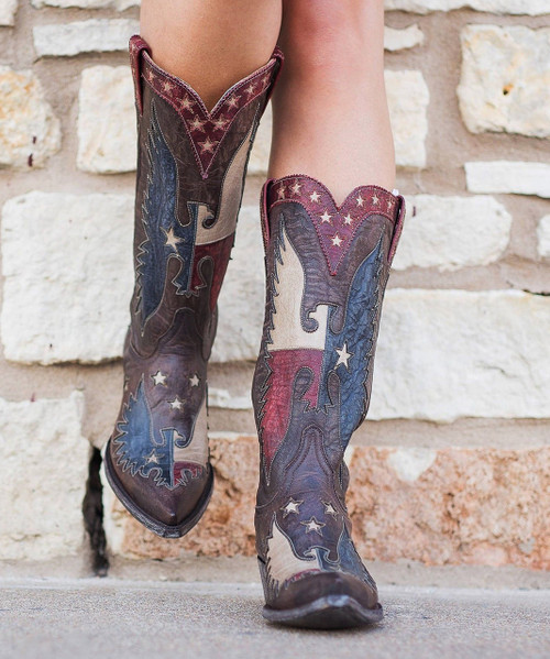 The boots are made from distressed brown leather and feature striking red, cream, and navy leather inlays forming large eagle wings and star motifs along the shaft and foot. The design resembles a stylized American flag theme, with scattered stars and sweeping wing shapes that give the boots a powerful, rugged look.

Additional details include:

Tall western shaft with red leather pull tabs decorated with embroidered stars

Pointed snip toe for a classic cowboy silhouette

Layered leather appliqué and contrast stitching

Distressed stacked heel and worn-in leather finish for a vintage character

The craftsmanship and artistic leatherwork give the boots a collectible western fashion feel, similar to statement styles often associated with artisan bootmakers like Old Gringo or Double D Ranch collaborations.

Overall, the boots project a bold, patriotic cowgirl style—perfect for someone who loves standout western footwear with handcrafted personality