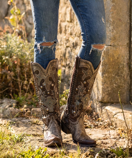 This is a **bold, highly embellished Western statement boot** that blends rugged cowgirl attitude with intricate Southwestern artistry. The rich **espresso-brown leather** is beautifully contrasted by **metallic brass and antique-gold accents**, creating a warm, aged, almost heirloom-inspired finish.

The shaft is covered in **ornate appliqué detailing, dimensional crosses, floral motifs, silver-toned studs, conchos, and hand-stitched embroidery**, giving the boot an incredible amount of texture and depth. The dramatic metallic brass overlays at the upper shaft and around the foot add a distinctive vintage-meets-rebel character.

One of the most eye-catching details is the **leather lace-up corset-style back**, which runs the length of the shaft and adds both visual interest and an adjustable element to the fit. The decorative side panels and embellished pull tabs continue the elaborate detailing from top to bottom.

The **snip toe and 1¾-inch western heel** give the boot a classic, flattering silhouette, while the combination of chocolate leather and metallic brass makes it surprisingly versatile—it would look incredible with denim, dresses, skirts, or layered boho pieces.

### Product description

**Double D Ranch Ammunition Boots – Metallic Brass & Espresso**

A true collector-worthy statement boot, the **Double D Ranch Ammunition** is where Western artistry meets rebellious cowgirl style. Rich espresso-brown leather is layered with shimmering metallic brass accents, intricate embroidery, dimensional crosses, ornate appliqués, antique-inspired studs, and dramatic Southwestern detailing.

The standout **corset-style leather lacing along the back of the shaft** adds an unexpected touch of attitude while complementing the boot’s beautifully embellished silhouette. Every inch is designed to make an impression, from the elaborate shaft detailing to the intricately decorated foot and classic snip toe.

These are not everyday Western boots—they're **wearable works of art** made for the woman who loves her boots to have personality, presence, and a little bit of rebellion. ?✨?

**Details:**

* **Shaft Height:** 16"
* **Shaft Opening:** 14"
* **Heel Height:** 1¾"
* **Toe:** Snip
* **Width:** Standard
* **Color:** Metallic Brass & Espresso Brown
* **Back:** Corset-style leather lacing
* **Style:** Western / Boho / Statement Boot
