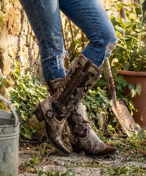 This is a **bold, highly embellished Western statement boot** that blends rugged cowgirl attitude with intricate Southwestern artistry. The rich **espresso-brown leather** is beautifully contrasted by **metallic brass and antique-gold accents**, creating a warm, aged, almost heirloom-inspired finish.

The shaft is covered in **ornate appliqué detailing, dimensional crosses, floral motifs, silver-toned studs, conchos, and hand-stitched embroidery**, giving the boot an incredible amount of texture and depth. The dramatic metallic brass overlays at the upper shaft and around the foot add a distinctive vintage-meets-rebel character.

One of the most eye-catching details is the **leather lace-up corset-style back**, which runs the length of the shaft and adds both visual interest and an adjustable element to the fit. The decorative side panels and embellished pull tabs continue the elaborate detailing from top to bottom.

The **snip toe and 1¾-inch western heel** give the boot a classic, flattering silhouette, while the combination of chocolate leather and metallic brass makes it surprisingly versatile—it would look incredible with denim, dresses, skirts, or layered boho pieces.

### Product description

**Double D Ranch Ammunition Boots – Metallic Brass & Espresso**

A true collector-worthy statement boot, the **Double D Ranch Ammunition** is where Western artistry meets rebellious cowgirl style. Rich espresso-brown leather is layered with shimmering metallic brass accents, intricate embroidery, dimensional crosses, ornate appliqués, antique-inspired studs, and dramatic Southwestern detailing.

The standout **corset-style leather lacing along the back of the shaft** adds an unexpected touch of attitude while complementing the boot’s beautifully embellished silhouette. Every inch is designed to make an impression, from the elaborate shaft detailing to the intricately decorated foot and classic snip toe.

These are not everyday Western boots—they're **wearable works of art** made for the woman who loves her boots to have personality, presence, and a little bit of rebellion. ?✨?

**Details:**

* **Shaft Height:** 16"
* **Shaft Opening:** 14"
* **Heel Height:** 1¾"
* **Toe:** Snip
* **Width:** Standard
* **Color:** Metallic Brass & Espresso Brown
* **Back:** Corset-style leather lacing
* **Style:** Western / Boho / Statement Boot
