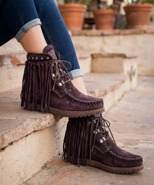 The El Vaquero ankle boot is a striking blend of bohemian spirit and rugged artisan craftsmanship. Handcrafted in Italy, this statement-making style features rich distressed suede in a deep plum-brown hue, highlighted by hand-stitched moccasin construction across the toe for an authentic, heritage-inspired look.

Long cascading fringe drapes dramatically from the ankle, creating beautiful movement with every step, while polished metal studs and vintage-inspired hardware add a touch of rock-and-roll edge. The lace-up front provides an adjustable fit, and the padded ankle cuff enhances both comfort and style.

Finished with a durable sole for everyday wear, this boot effortlessly combines Western, Native-inspired, and boho-chic influences into a truly distinctive silhouette. Pair it with denim, flowing dresses, or layered festival looks for an effortlessly free-spirited statement.

Features:
• Handmade in Italy
• Distressed suede leather upper
• Hand-stitched moccasin toe construction
• Dramatic fringe detailing
• Studded ankle cuff accents
• Lace-up front closure
• Antiqued metal hardware
• Durable lug outsole
• Signature El Vaquero artisan craftsmanship