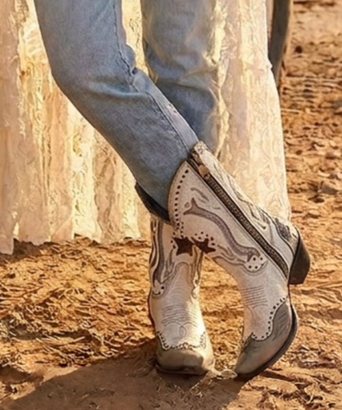 These boots are a striking blend of vintage Western tradition and Americana-inspired artistry. With their weathered finish, handcrafted details, and iconic lucky symbols, they feel like a pair that has been collected over decades and passed down with stories to tell.

Product Description

Channel timeless Western heritage with these statement-making handcrafted boots that celebrate luck, freedom, and authentic cowboy style. Crafted from beautifully distressed crackled ivory leather, they feature a perfectly broken-in vintage finish that gives each pair incredible character from the very first wear.

The tall shaft is adorned with layered leather appliqués, whipstitched trim, antique nailhead studs, and intricate embroidery that create remarkable depth and texture. At the center of the design, a classic horseshoe and Western star symbolize good fortune and adventure, while sweeping gray leather inlays add movement inspired by traditional cowboy scrollwork.

The soft gray distressed toe and heel counters create a beautiful contrast against the creamy white leather, enhancing the boot's rustic appeal. Antique-inspired studding outlines every panel, giving the design an heirloom quality reminiscent of vintage rodeo boots and collectible Western pieces.

A square snip toe, leather sole, and stacked cowboy heel complete the silhouette with comfort and timeless style, while the hand-finished distressed leather ensures no two pairs are exactly alike.