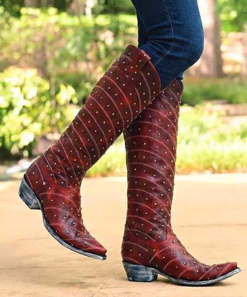 Make a statement in every step with these bold, handcrafted western boots—where classic cowgirl style meets elevated, artisan detail.

Crafted from premium black leather, these boots feature intricate contrast stitching in a feather-inspired pattern that flows across the shaft and vamp, delivering timeless western character with a refined edge. The sleek pointed toe and angled heel create a flattering, feminine silhouette that’s both versatile and striking.

What truly sets this pair apart is the ornate vertical overlay panel, adorned with hand-set metal studs and rich red stone accents. A signature Double D Ranch medallion charm drapes elegantly from the panel, adding movement and a collectible, heritage-inspired touch.

At the back, a contrasting textured leather heel counter with a spotted, exotic finish brings unexpected dimension and standout style. A side zipper ensures easy on-and-off without compromising the clean, classic profile.

Perfect for pairing with dresses, denim, or statement looks, these boots embody western tradition with a bold, fashion-forward spirit.

Details:

Premium black leather construction

Intricate contrast western stitching

Studded overlay with red stone embellishments

Signature medallion charm detail

Textured heel counter for added contrast

Side zipper closure

Pointed toe & stacked heel

Confident. Detailed. Unmistakably cowgirl.