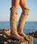 Close-up fashion editorial shot of a pair of tall boho western boots in deep dusty rose suede leather, worn by a model standing on coastal rocks near the ocean. The boots are mid-thigh height with a relaxed slouchy fit, featuring long flowing fringe along the outer seams, decorative whipstitch detailing, metal ring hardware, braided trim around the top edge, and a star patch medallion with tassel accent near the knee. The soles are thick rubber with geometric western stitching. The model’s bare legs are visible above the boots. Background shows a soft blue ocean horizon under clear sky with warm golden sunlight creating rich shadows and a dreamy western coastal mood.