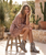 This image captures a sun-soaked, feminine western-boho vibe with soft, romantic tones ?✨

A woman sits casually on a rustic wooden stool outdoors, surrounded by desert-style greenery and warm stone textures. The setting feels relaxed and earthy, with natural light casting a golden glow that enhances the soft, dreamy aesthetic.

She wears a flowy, off-the-shoulder mini dress in a muted blush-brown tone, featuring delicate patterns and airy sleeves that add movement and a touch of vintage romance.

Her look is beautifully coordinated:

A light pink cowboy hat adds a playful yet polished western touch
Matching blush suede ankle boots stand out with intricate details like fringe, moccasin-style stitching, and decorative trim
The boots also feature subtle metallic accents, tying in a handcrafted, elevated feel

Her relaxed pose—resting her head on her hand with a soft smile—gives the image an effortless, approachable charm.

Overall, the look blends soft femininity with western edge, creating a style that feels both modern and timeless, perfect for warm days with a hint of rustic elegance