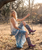 The photo shows a soft, romantic boho-western scene set outdoors in a rustic countryside yard with trees, dry grass, and warm natural sunlight.

A young woman sits sideways on a simple wooden rope swing, gently holding the rope with one hand and looking toward the camera with a calm, thoughtful expression. Her hair is styled naturally in a loose updo, and her makeup is minimal, giving her a fresh, effortless look.

She’s wearing a flowy bohemian outfit—a delicate cream lace vest layered over a long blue paisley-print maxi skirt. The fabrics move softly and add to the relaxed, free-spirited feel of the scene.

The standout piece is her tall cowgirl boots, made of light tan leather with hand-painted-style heart designs scattered across the shaft. The boots have a classic western silhouette with a pointed snip toe and stacked heel, adding a playful, feminine twist to the outfit.

Overall mood: romantic, vintage-inspired, and peaceful, blending country charm with boho fashion and highlighting the boots as a whimsical, eye-catching detail.