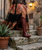 This image embodies dark romantic bohemian luxury with a touch of Western sophistication.

The model is descending weathered stone steps wearing a dramatic floor-length dress featuring a rich floral print in shades of coral, rose, sage, gold, teal, and blush layered beneath intricate black lace. The plunging neckline, fitted waist, and flowing skirt create a striking silhouette that feels both feminine and effortless.

A high front slit reveals a pair of sleek black knee-high leather boots, adding an unexpected edge to the romantic dress and grounding the look with confidence and sophistication. Delicate layered jewelry and softly tousled hair complete the free-spirited, worldly aesthetic.

The backdrop of aged stone and pastel architecture enhances the feeling of old-world charm and wanderlust, creating a look that feels equally at home in: