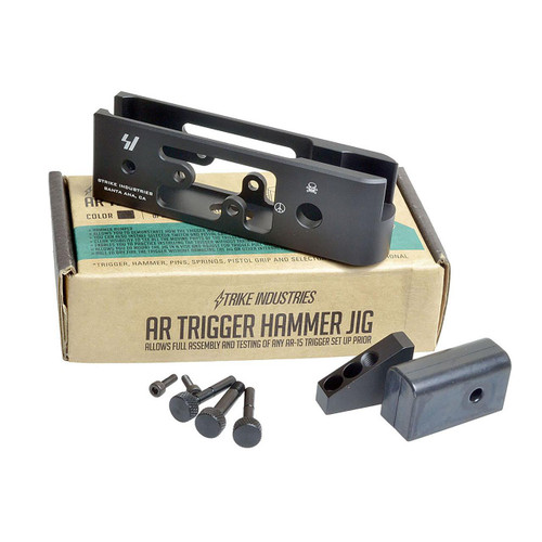 Strike Industries SI-AR-THJ AR Trigger Hammer Jig, a black metal tool for firearm trigger and hammer assembly testing, placed on top of its packaging box with additional small parts and screws around it.