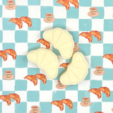 A box of Blackcurrant & Sweet Vanilla scented wax melts in the shape of croissants, shown inside their illustrated gift box and resting beside it. The FSC-certified packaging features a hand-painted tabletop scene by Sarah Pettitt including a croissant, coffee pot and berries on a pink chequered tablecloth.