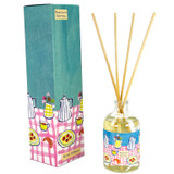 A Heaven Scent 100ml clear apothecary glass reed diffuser with four natural reeds, standing next to its tall teal-blue gift box. The box and bottle label feature a vibrant illustration by Sarah Pettitt of a pink and white chequered table set with blue striped coffee pots, yellow chequered jugs with flowers, croissants and plates of red berries.