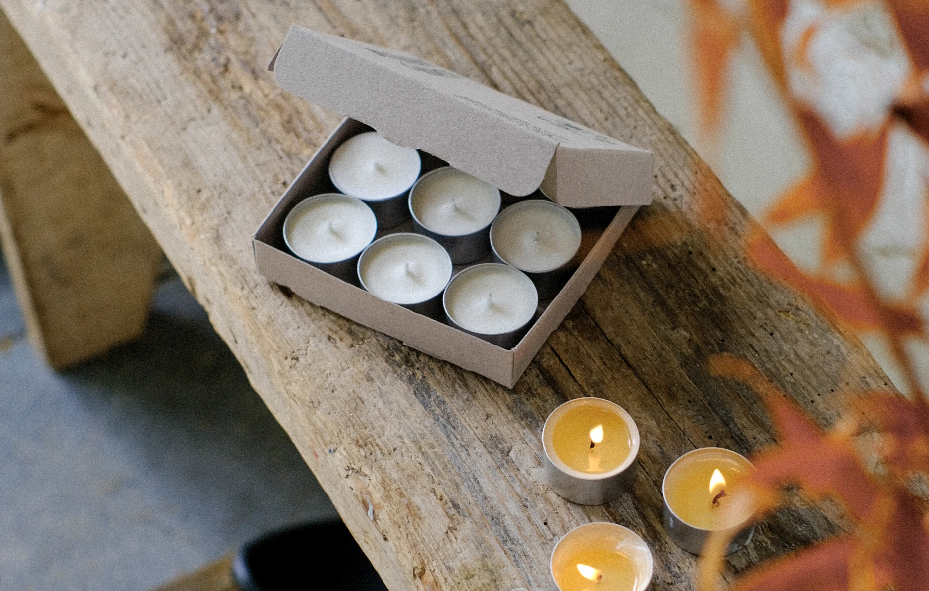 An open box of nine natural soy wax tealights and several lit tealights displayed on a weathered wooden plank alongside autumn foliage.