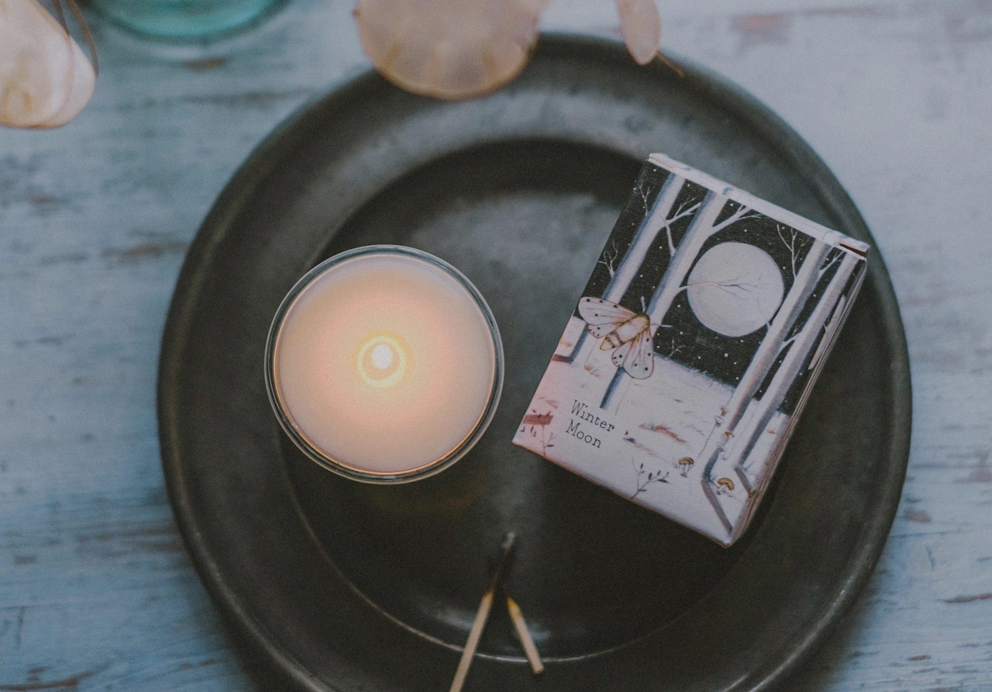 A lit 9cl Winter Moon scented votive candle in clear glass alongside its illustrated box, displayed on a pewter tray beneath delicate dried botanical stems.