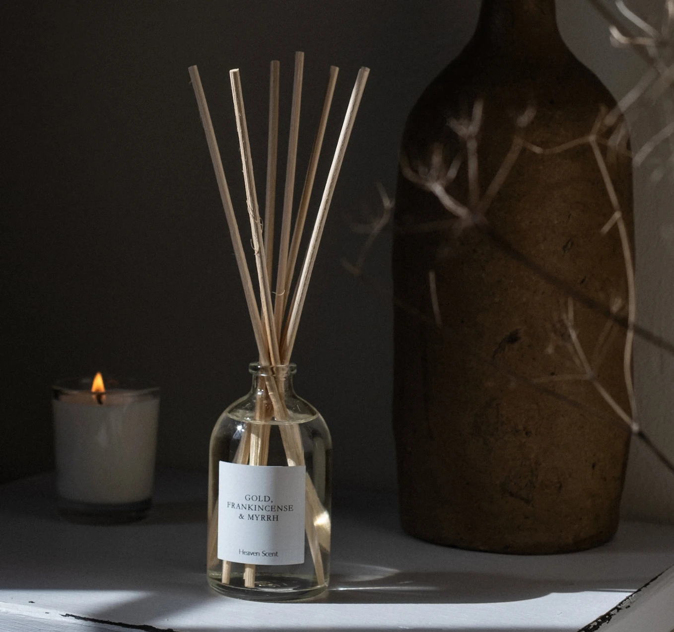 A 100ml Gold, Frankincense & Myrrh apothecary reed diffuser in a clear glass bottle with natural reeds, displayed alongside a lit candle and a rustic ceramic container.