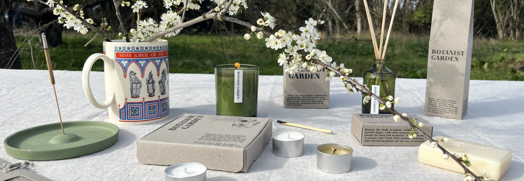 Heaven Scent's Green Botanical range, showing the Botanist Garden Collection on a summer table with apple blossom. Lit candles, reed diffuser, incense and soap all look beautiful in a sunny setting