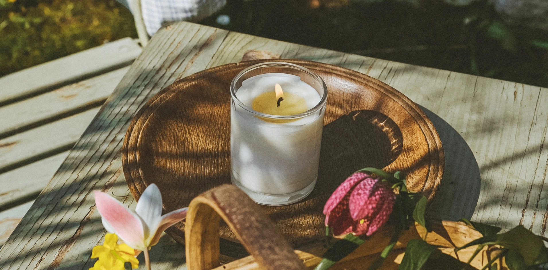 A lit summer natural soy vegan candle in clear glass resting on a wooden tray with spring flowers.