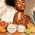 Black woman gently applying Citrus + Agave Body Butter to her shoulder beside the orange-and-gold labeled jar and citrus fruit.