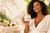 Black woman holding Lime + Coconut Body Butter with its green-and-gold label in a bright editorial lifestyle scene.