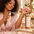 Woman lightly misting Rose Nectar Perfume toward her inner wrist beside the matching labeled kraft tube.