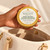 Hand holding the palm-sized 4oz Gold Tin Body Butter above an ivory handbag; Serenity Green Tea is shown.