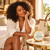 Black woman enjoying a quiet hotel-room moment beside her handbag and current-label 4oz Gold Tin Body Butter; Serenity Green Tea is shown.