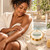 Black woman gently applying Unscented Body Butter to her forearm in a sunlit bath setting.