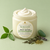 Serenity Green Tea Body Butter with its pale-green label and gold Dame mark beside tea leaves and stones.