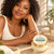 A Black woman in an ivory knit top rests her hand on her forearm beside Nourishing Eczema Body Butter.