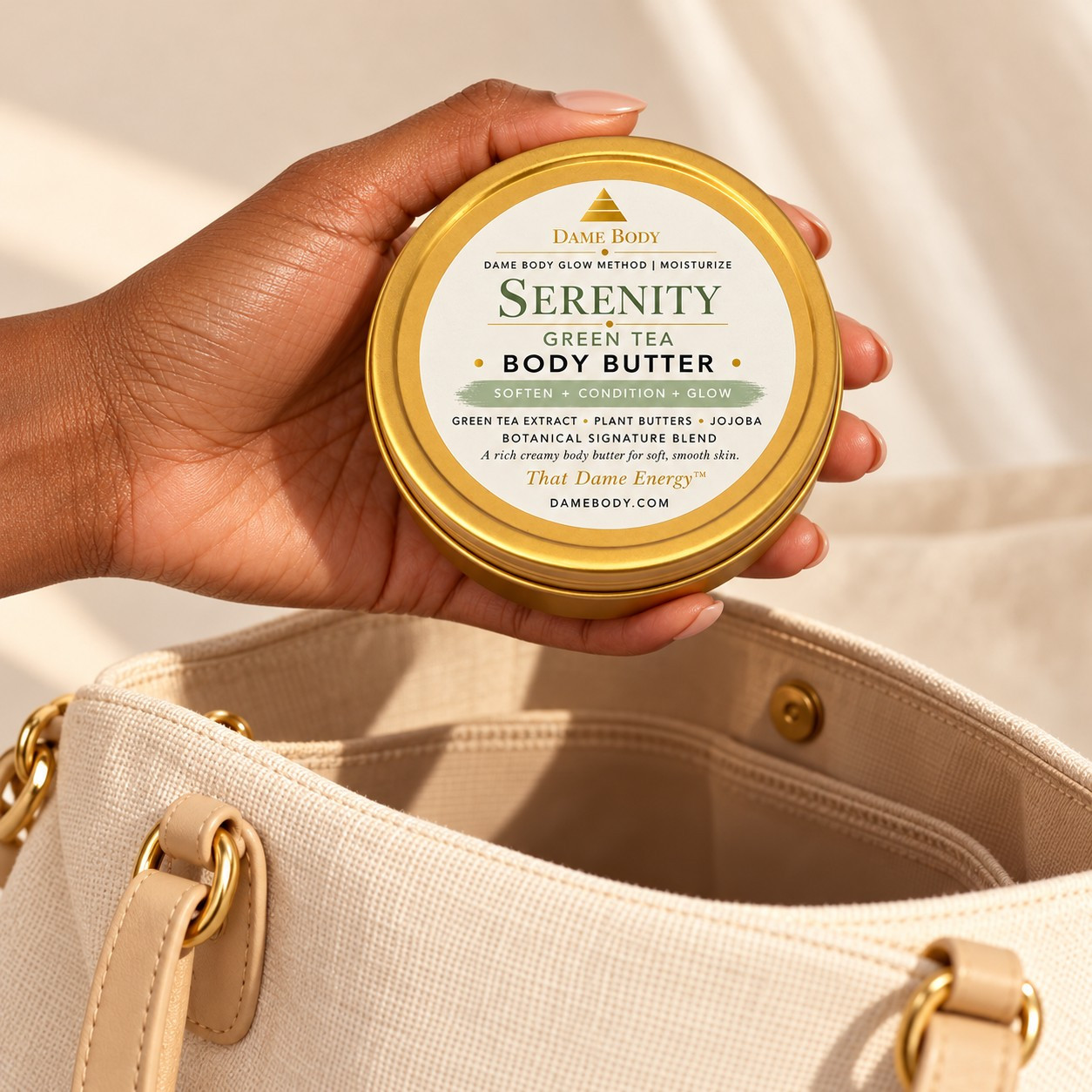 Hand holding the palm-sized 4oz Gold Tin Body Butter above an ivory handbag; Serenity Green Tea is shown.
