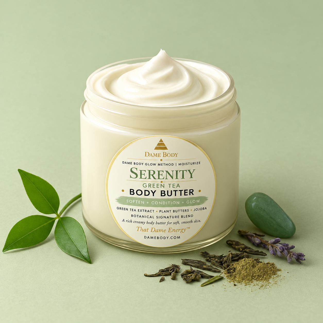 Serenity Green Tea Body Butter with its pale-green label and gold Dame mark beside tea leaves and stones.