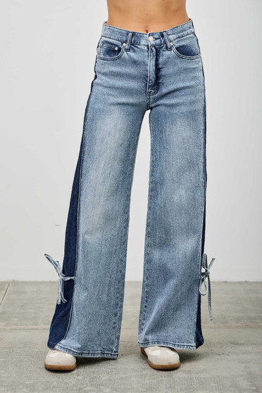 "A front view of a person wearing high-waisted, wide-leg denim jeans. The jeans feature a two-tone design with a light-wash front and dark indigo side panels. At the outer ankles, there are small slits tied with denim bows. The person is also wearing white sneakers, standing against a neutral grey background."