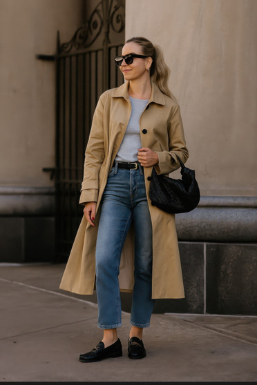 "Woman wearing a tan single-breasted trench coat with straight-leg blue jeans, black loafers, and a black woven handbag in a street style fashion setting."