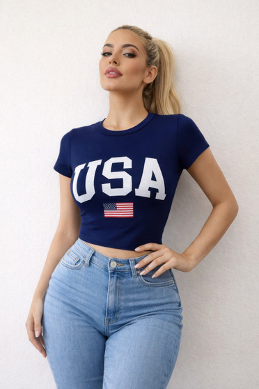 "A blonde woman in a fitted navy blue cropped t-shirt featuring a white 'USA' graphic and an American flag icon, paired with high-waisted light wash denim jeans against a white textured wall."