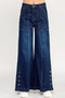 Front view of a person wearing high-waisted, dark wash wide-leg denim jeans. The jeans feature prominent vertical seams down the center of each leg, large front patch pockets, and a row of decorative metallic buttons at the outer hem of the flared legs.