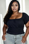 Plus-size woman wearing a black short-sleeve smocked waist top with puff sleeves and grey jeans.
