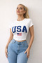 Woman wearing a white "USA" graphic baby tee with an American flag print and high-waisted light wash jeans against a white textured wall.