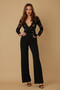 Model wearing a black long-sleeve jumpsuit featuring a floral lace scalloped V-neck bodice, a belted high waist with a gold buckle, and tailored wide-leg trousers.