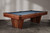 Three-quarter angle view of the Doc & Holliday Preakness pool table, showing the walnut finish, maple racing stripe detail, and pedestal base design.