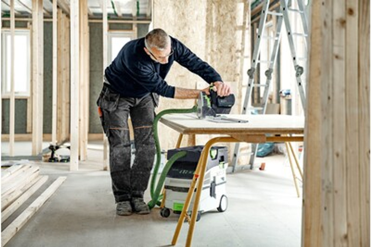 The image depicts a construction worker performing a task in a construction zone. 
Foreground:
o	The worker is operating a Festool plunge-cut saw on a wooden panel placed on a makeshift worktable.
o	A Festool dust extractor is connected to the saw via a green hose, ensuring dust collection during the cutting process.
o	The saw and extractor are professional-grade tools, designed for precision and cleanliness.
Worker:
o	The individual is wearing dark work attire, including a navy sweater and gray pants with utility pockets and reinforced knees.
o	Protective eyewear is worn, demonstrating adherence to safety protocols.