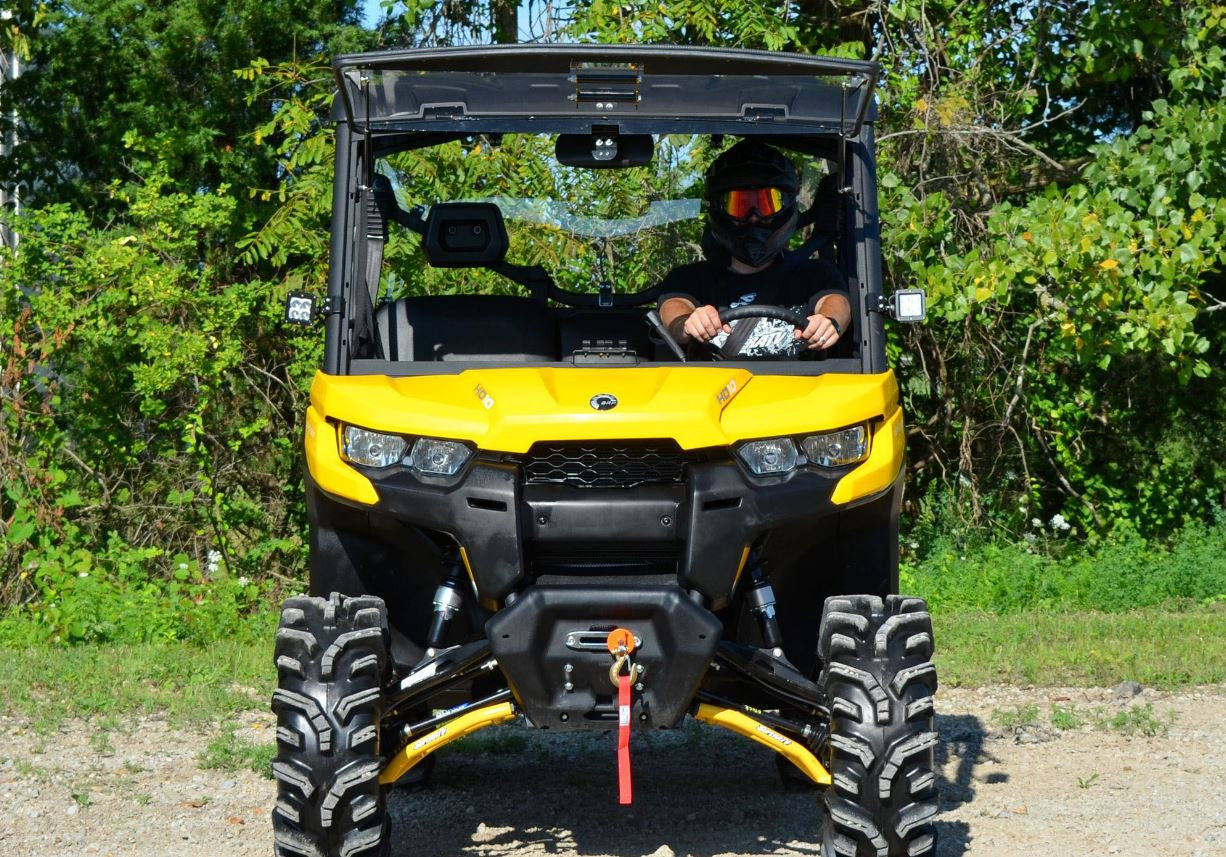 Straight-on front view of the yellow Can Am Defender with the SuperATV windshield closed and driver inside
