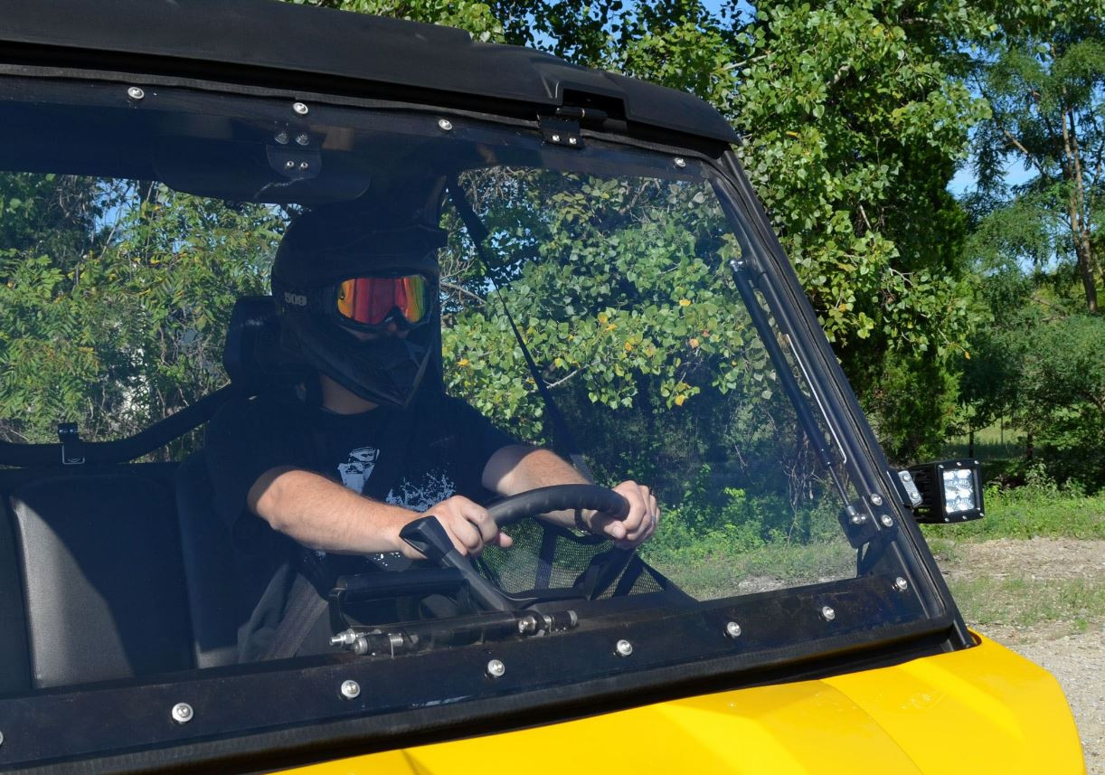 Straight-on close-up of the closed SuperATV windshield with the helmeted driver behind it on the Can Am Defender