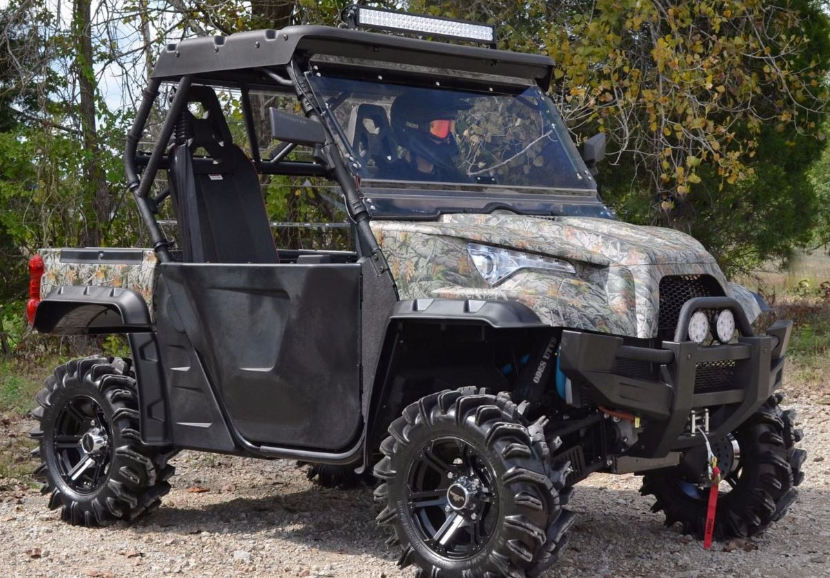 Full side view of a camo Odes Dominator with the SuperATV flip-up windshield, parked on gravel with mud tires