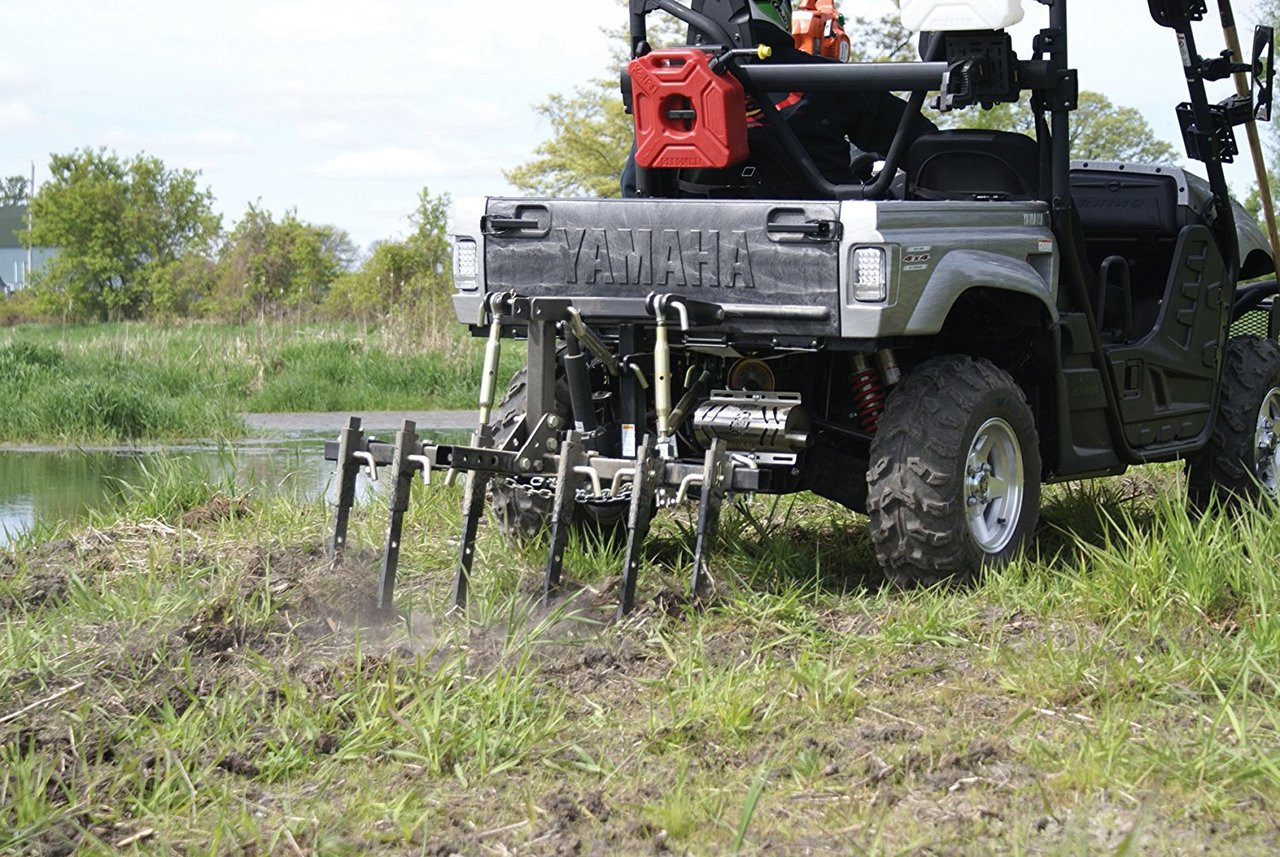 Kolpin Dirtworks 3 point hitch with cultivator tines working grassy ground behind a gray UTV by a pond