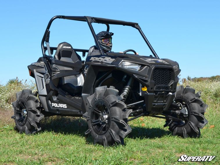 Black side-by-side fitted with four SuperATV Assassinator mud tires, front three-quarter view in a field