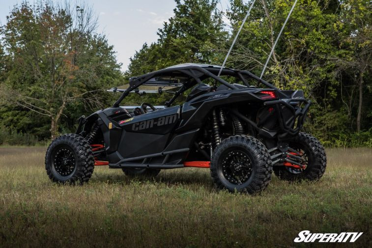 Front three-quarter view of the black Maverick X3 with SuperATV nerf bars, parked on grass with trees behind