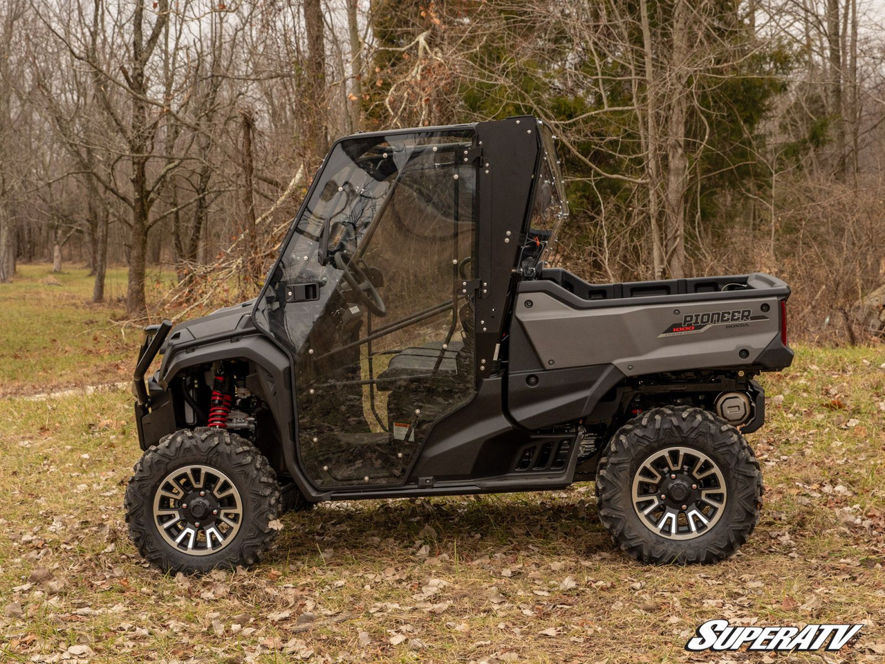 SuperATV full cab enclosure doors installed on a gray Honda Pioneer 1000, driver side view in the woods