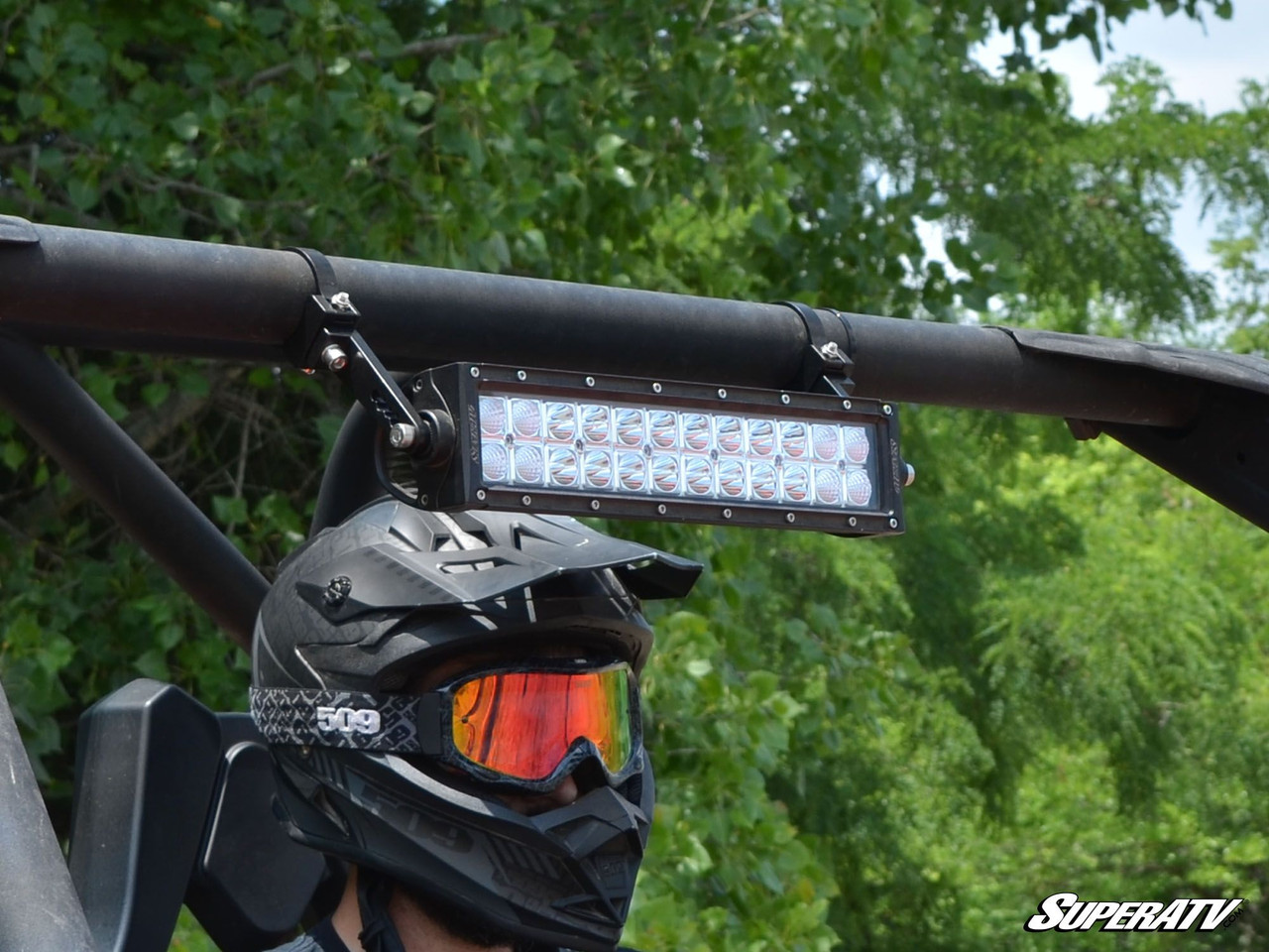 LED light bar held by SuperATV aluminum tube clamps on a roll cage above a driver in a black helmet and goggles