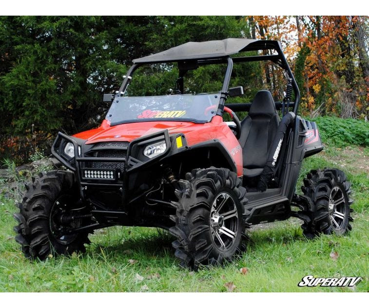 Front three-quarter view of a red Polaris RZR on mud tires with the SuperATV rock sliding nerf bars, in the woods