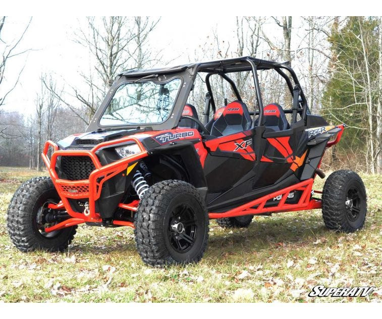 Front three-quarter view of a red four-seat Polaris RZR XP with SuperATV nerf bars installed, parked in the woods
