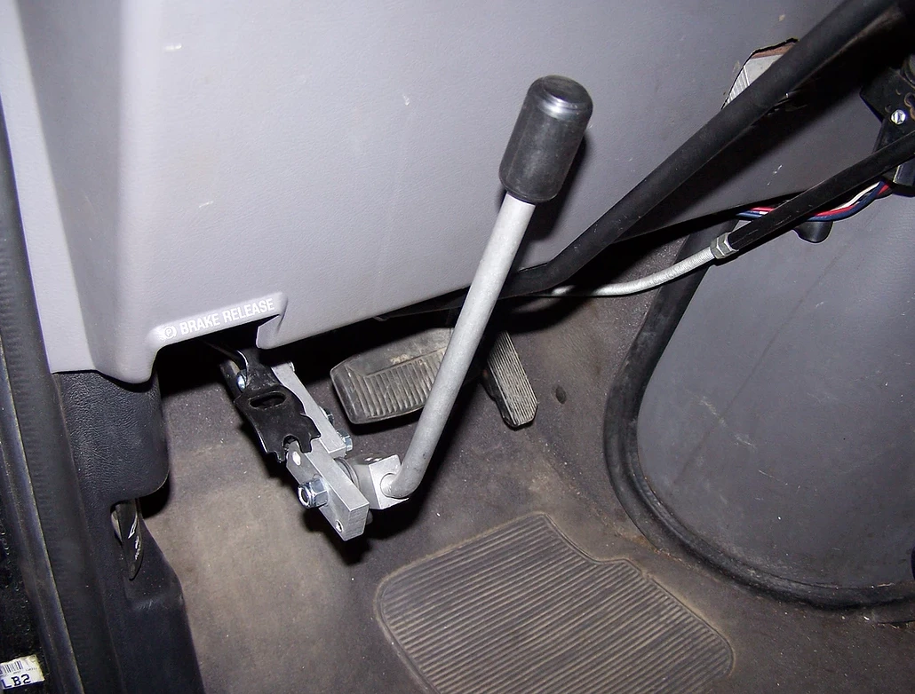 Sure Grip park brake extension lever installed on a vehicle floor next to the pedals, angled view