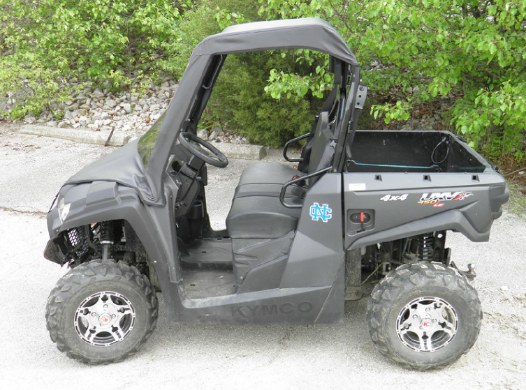 Side view of a gray Kymco 450 with the 3 Star soft top and vinyl windshield, parked on gravel
