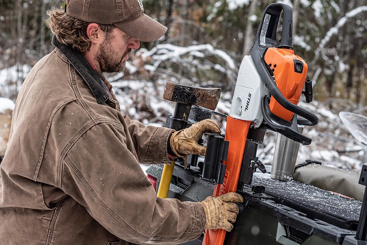 Man in a brown jacket lifting a chainsaw out of the Kolpin chainsaw mount on a side-by-side in a snowy forest